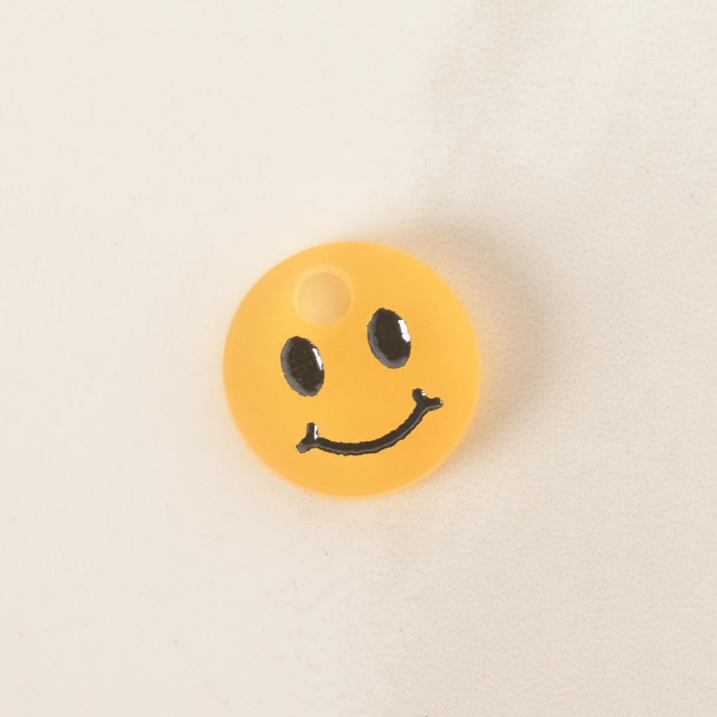 A close-up of a small, translucent yellow, circular pet tag charm with a black smiley face. The charm is displayed on a light-colored, speckled countertop or stone surface.