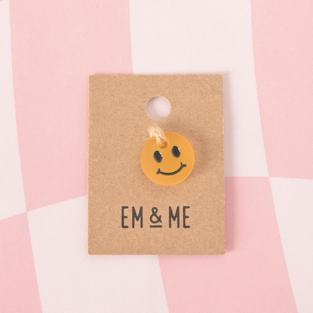 A small, translucent orange, circular pet tag charm with a black smiley face. The charm is attached with a piece of string to a brown cardboard tag with the logo "EM & ME." The item is displayed on a pink and white checkered background.