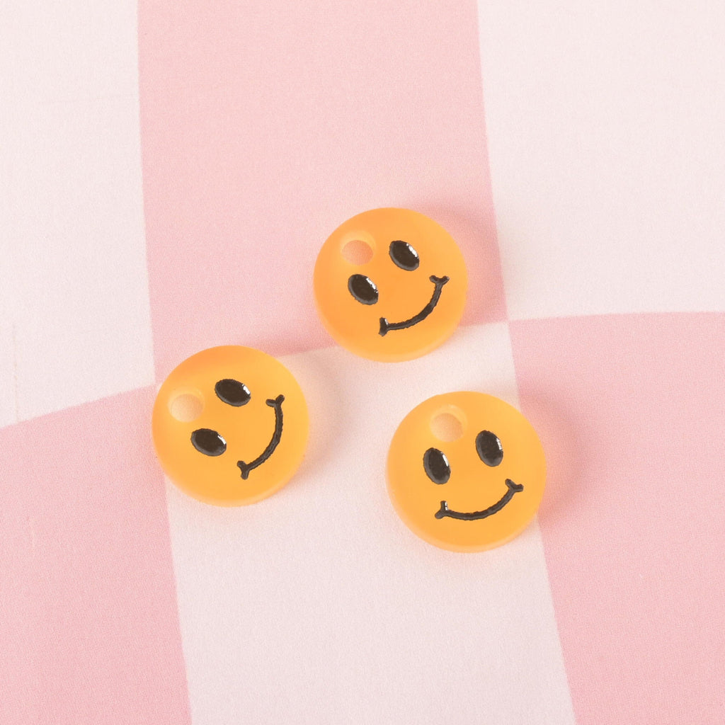 A group of three small, translucent Yellow, circular pet tag charms with black smiley faces. The charms are displayed on a pink and white checkered background.