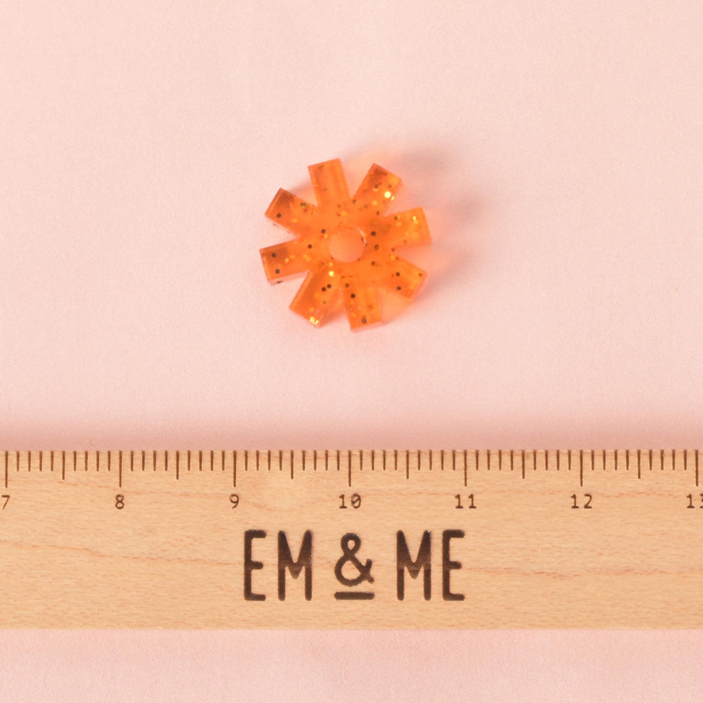 A small, glittery orange, starburst-shaped pet tag charm. The charm is displayed above a wooden ruler with the logo "EM & ME." The background is a solid pink.