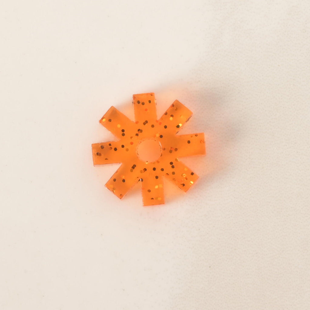 A close-up of a small, translucent orange, starburst-shaped pet tag charm. The charm has a subtle glitter finish with black specks. It is displayed on a light-colored, speckled countertop or stone surface.
