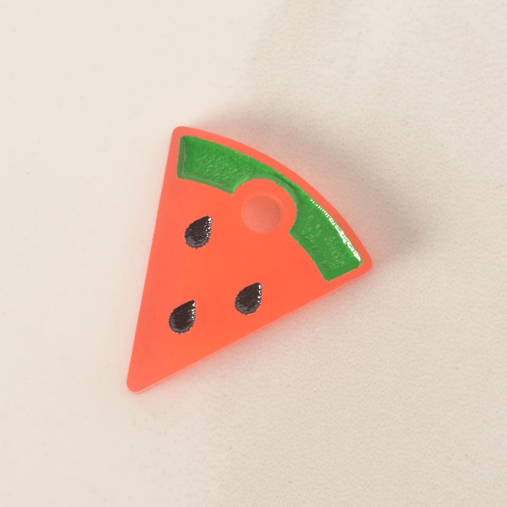 A close-up of a small, pink and green watermelon slice-shaped pet tag charm with three black seeds. The charm is displayed on a light-colored, speckled countertop or stone surface.