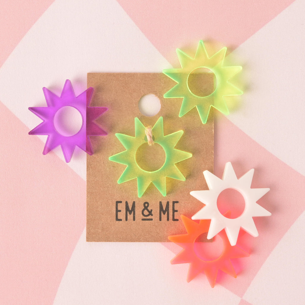 A collection of six starburst-shaped charms or pins in translucent neon colors: purple, yellow-green, white, and neon orange. One yellow-green charm is attached to a brown paper card that reads "EM & ME." The items are displayed on a pink and white checkered background.