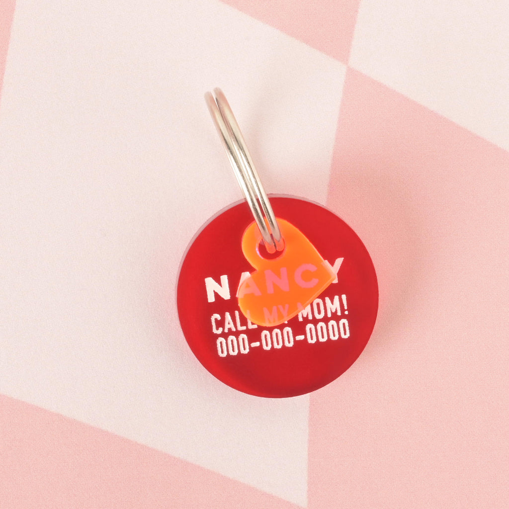 A close-up product photo of a round, red pet tag, also known as a Minimal Circle tag, with a neon orange heart charm attached to the top. The tag is engraved with the name "NANCY" and the funny phrase "CALL MY MOM!" It is shown with a silver split key ring.