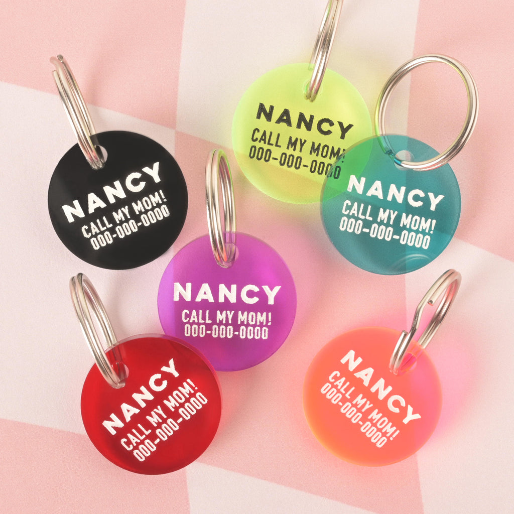 A product photo showing six round, custom pet ID tags in various colors. From left to right, the tags are: black, neon yellow, teal, purple, red, and a red-to-orange gradient. All tags are engraved with the name "Nancy" and the phrase "Call My Mom!" and are shown with a silver split key ring.