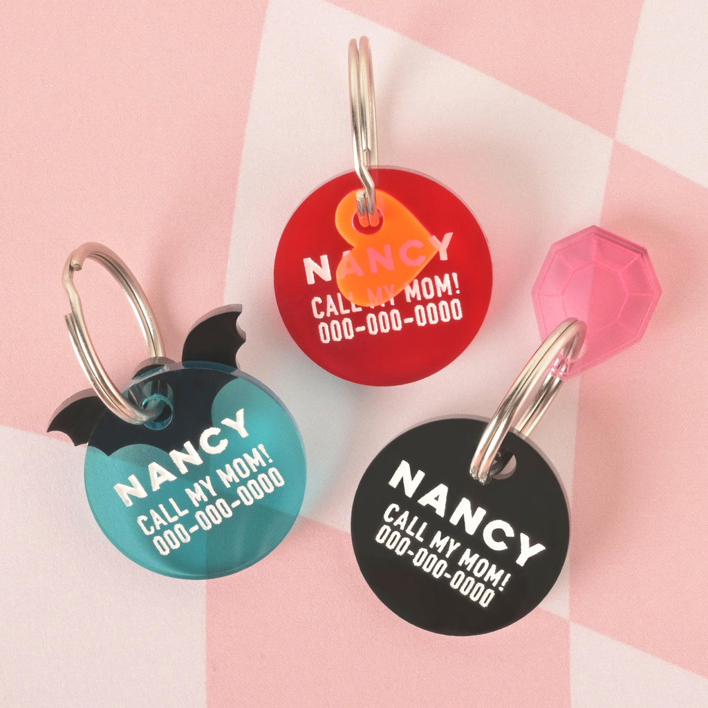 A product photo showing three round pet ID tags with a "Call My Mom!" engraving. The top tag is red with a neon orange heart charm. The bottom left tag is teal with black bat wings. The bottom right tag is black with a translucent pink gemstone charm. All three tags are engraved with the name "NANCY" and have a silver split key ring.