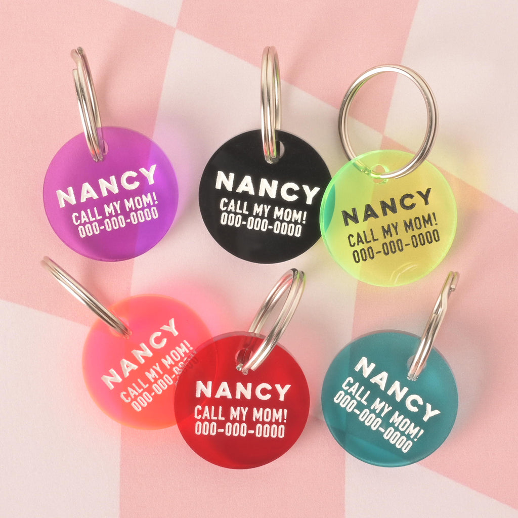 A product photo showing six round, custom pet ID tags in various colors on a checkered pink and white background. The tags are purple, black, neon yellow, a red and orange gradient, red, and teal. All tags are engraved with the name "NANCY" and the funny phrase "CALL MY MOM!" and are shown with a silver split key ring.