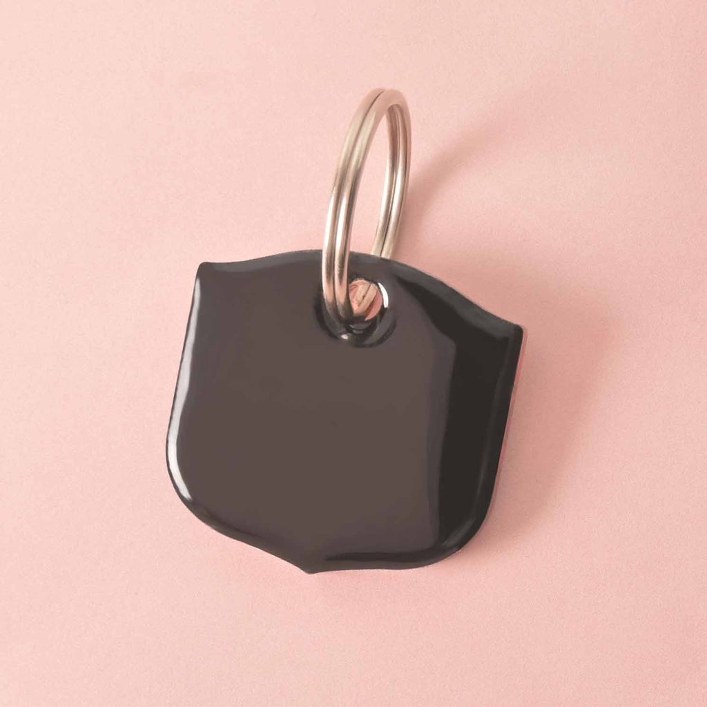 A blank, black shield-shaped pet tag attached to a silver ring, perfect for a collar. The tag has a shiny surface and is resting on a solid pink background.