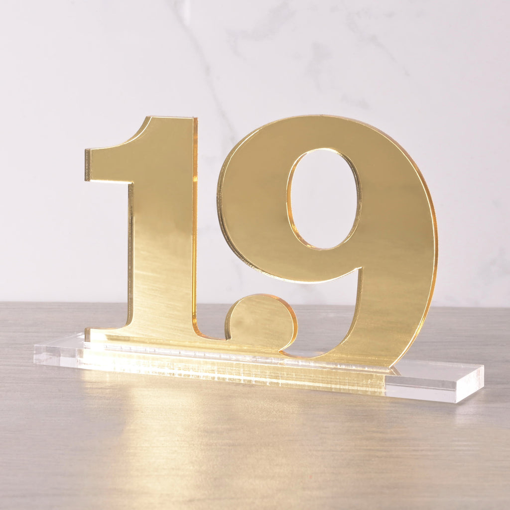 Gold acrylic table number “19” with a sleek clear base, displayed on a wooden surface for weddings or events.