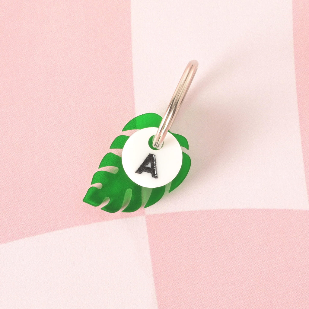 A custom, dark green, monstera leaf-shaped pet tag. A small, white, circular charm with a black letter "A" is attached to the shiny silver keyring, sitting in front of the leaf charm. The tags are displayed on a pink and white checkered background.