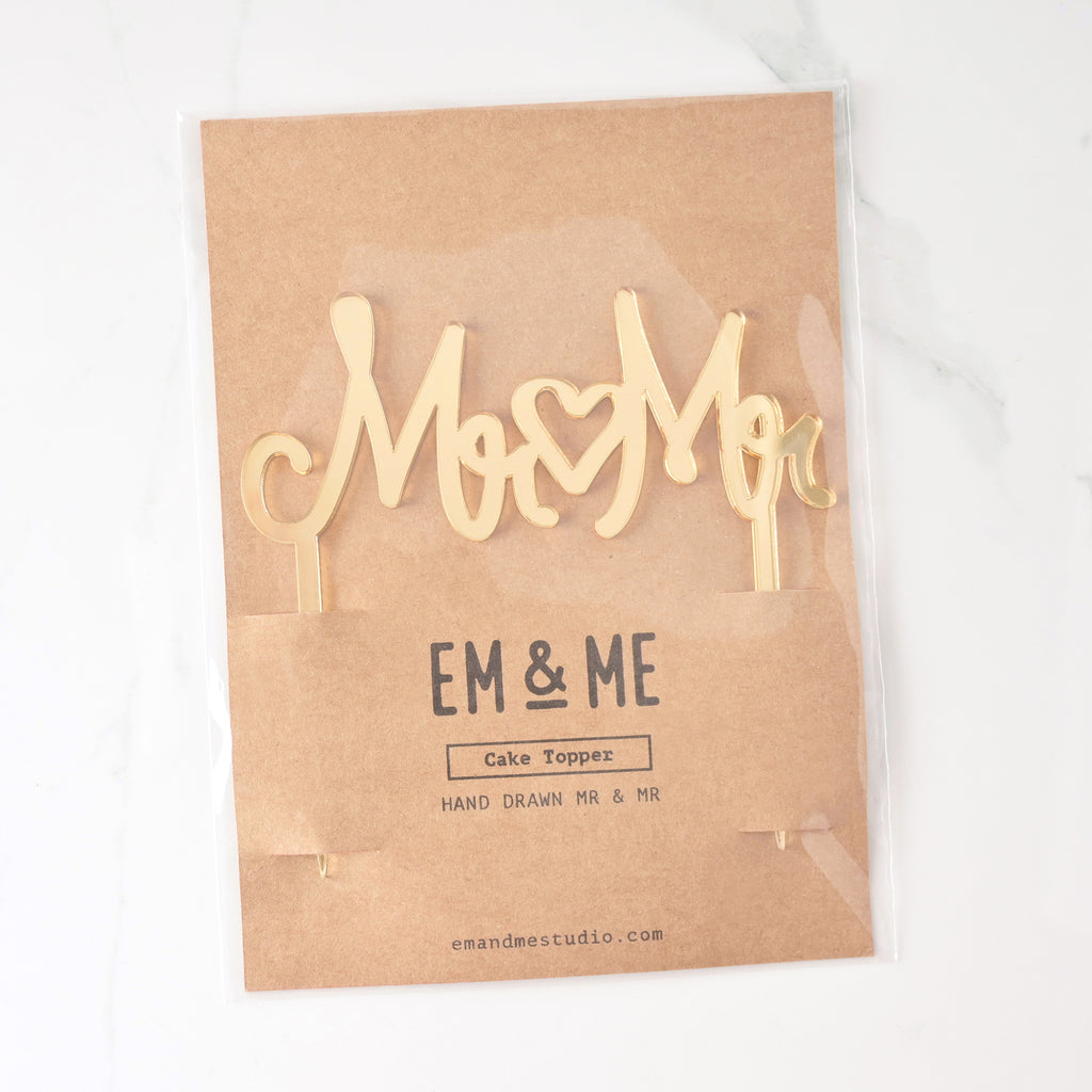 mirror gold cake topper that reads "MR MR" with a heart in the middle for wedding Packaged on brown kraft paper. Inclusive LGBTQIA+ celebration.