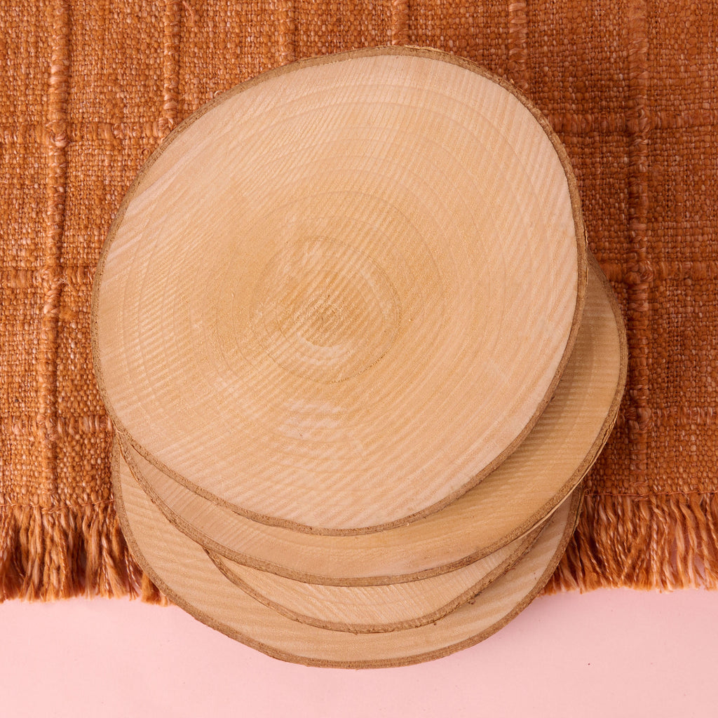 A stack of four circular, natural wood slices with visible growth rings and bark edges, perfect for use as rustic coasters or craft blanks. They are resting on a rust-colored woven textile.