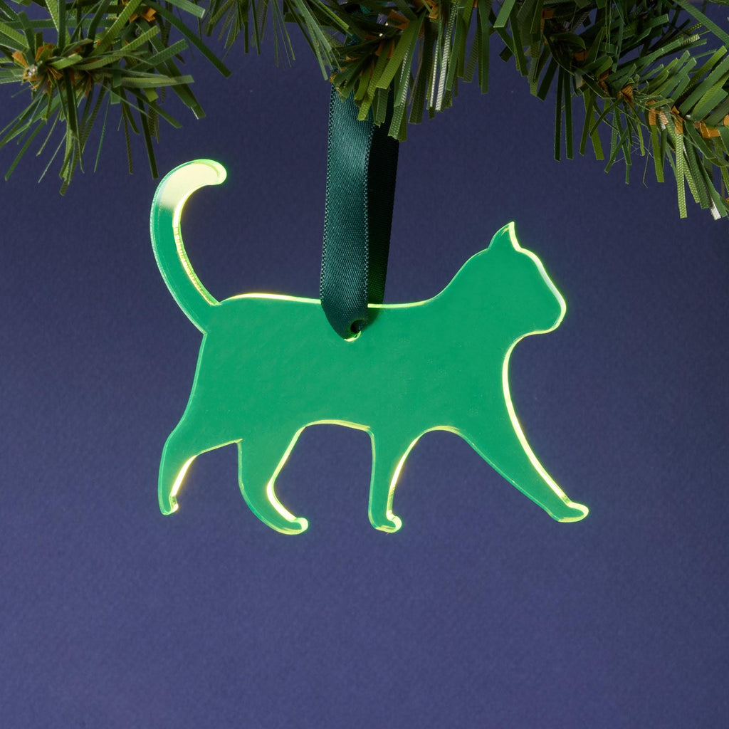 A laser-cut fluorescent neon green acrylic cat silhouette ornament, hanging by a dark green ribbon from a pine branch. This vibrant pet décor is set against a dark blue background.