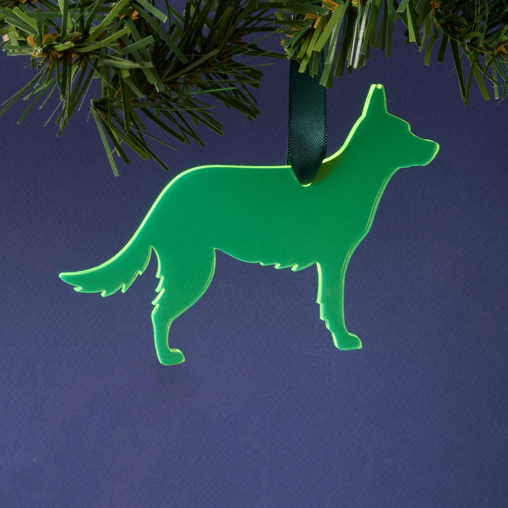 A laser-cut German Shepherd silhouette ornament made from fluorescent neon green acrylic, hanging by a dark green ribbon from a pine branch. This vibrant pet décor is set against a dark blue background.