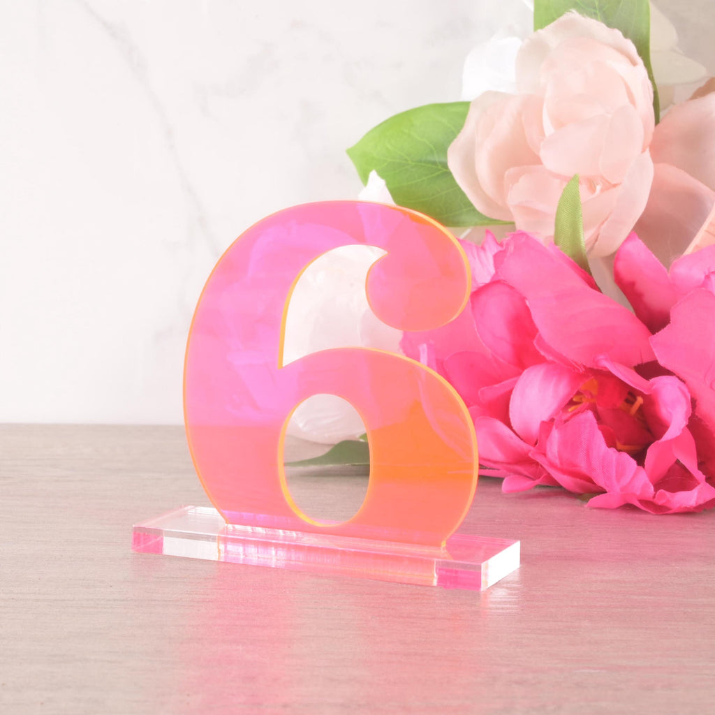 Neon pink acrylic table number “6” on a clear base, displayed with pink and blush flowers, perfect for wedding or party centerpieces.