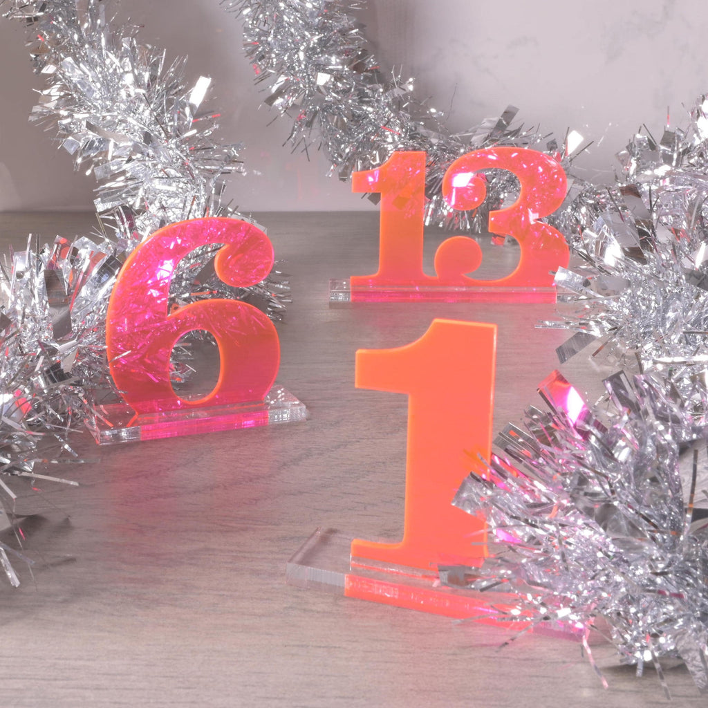 Bright neon pink acrylic table numbers “1,” “6,” and “13” on clear bases, styled with silver tinsel garland for festive wedding or party decoration.