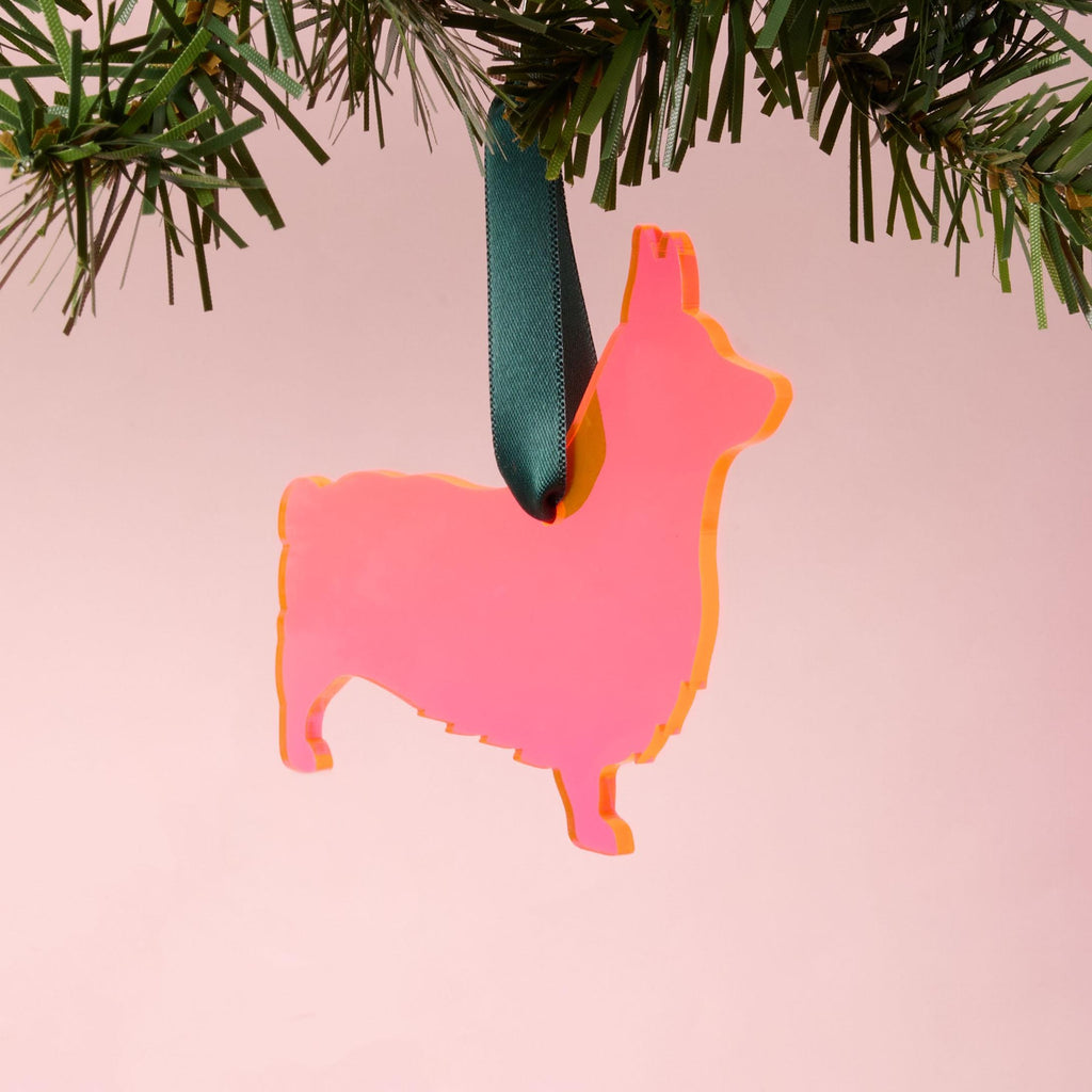 A laser-cut Corgi silhouette ornament made from vibrant fluorescent pink and orange acrylic, hanging by a dark green ribbon from a pine branch, set against a pink background.