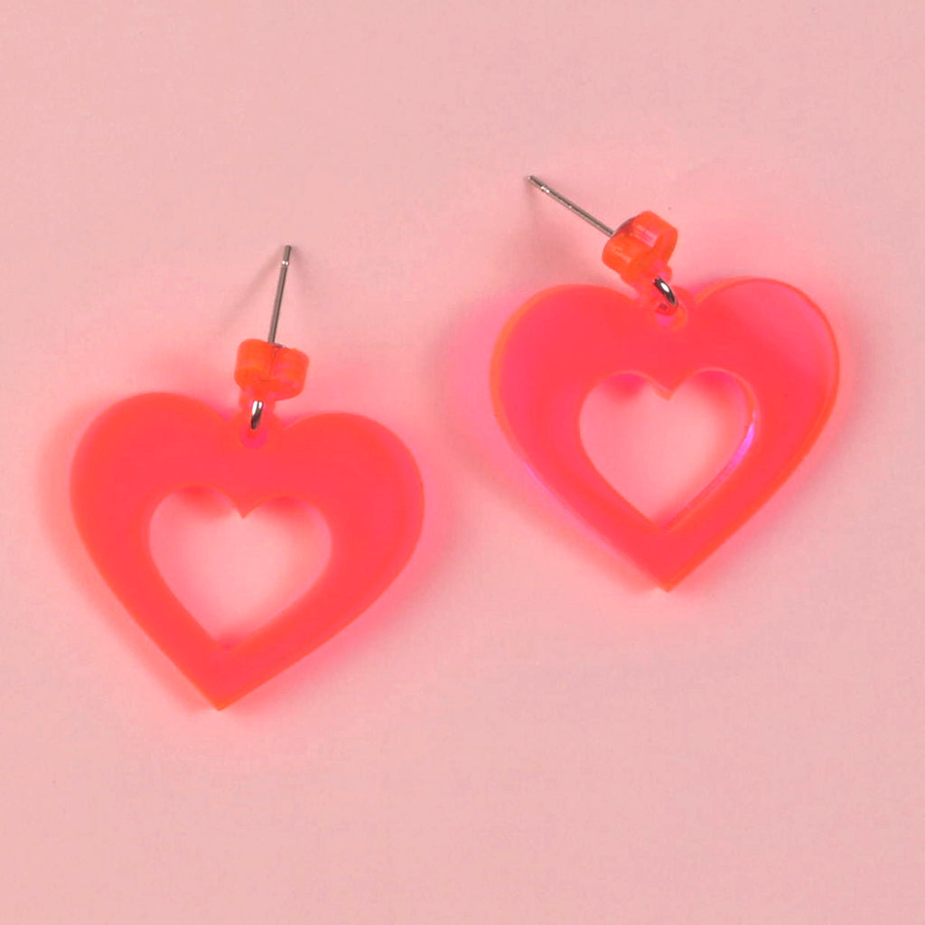 A pair of large, laser-cut, bright fluorescent pink acrylic heart-shaped cutout earrings with orange stud posts, displayed against a soft pink background.