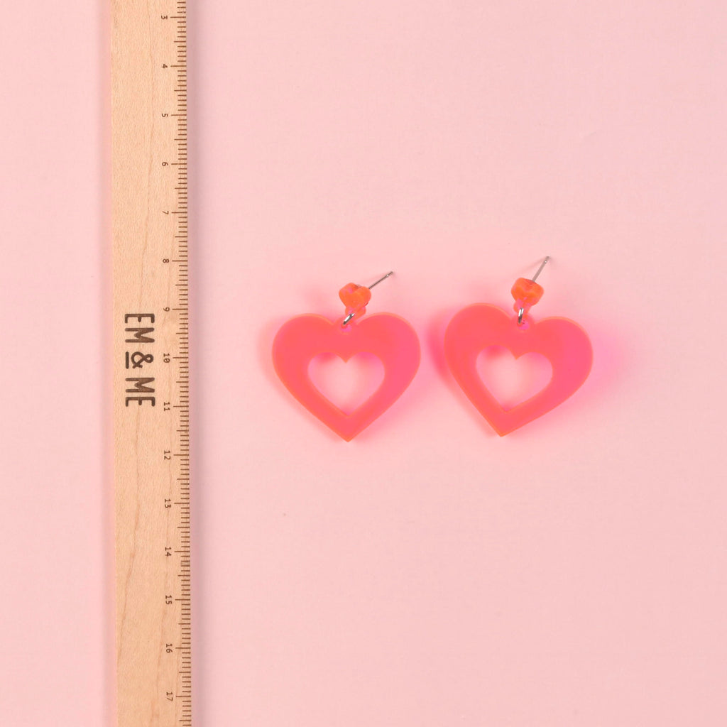 A pair of large, bright fluorescent pink acrylic heart-shaped cutout dangle earrings with orange studs. A wooden ruler is shown on the left to indicate the size of the jewelry.
