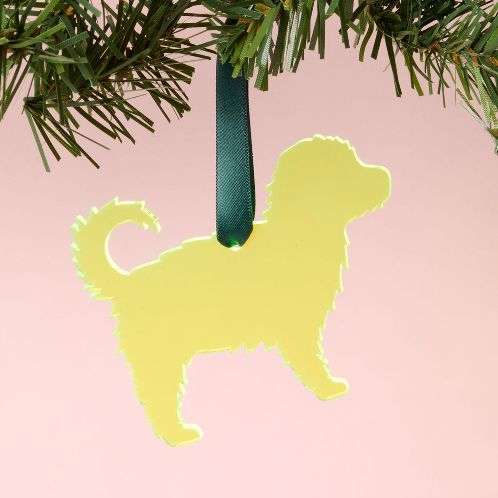 A laser-cut fluffy dog silhouette ornament (such as a Goldendoodle or Poodle Mix) made from fluorescent neon yellow-green acrylic, hanging by a dark green ribbon from a pine branch, set against a pink background.