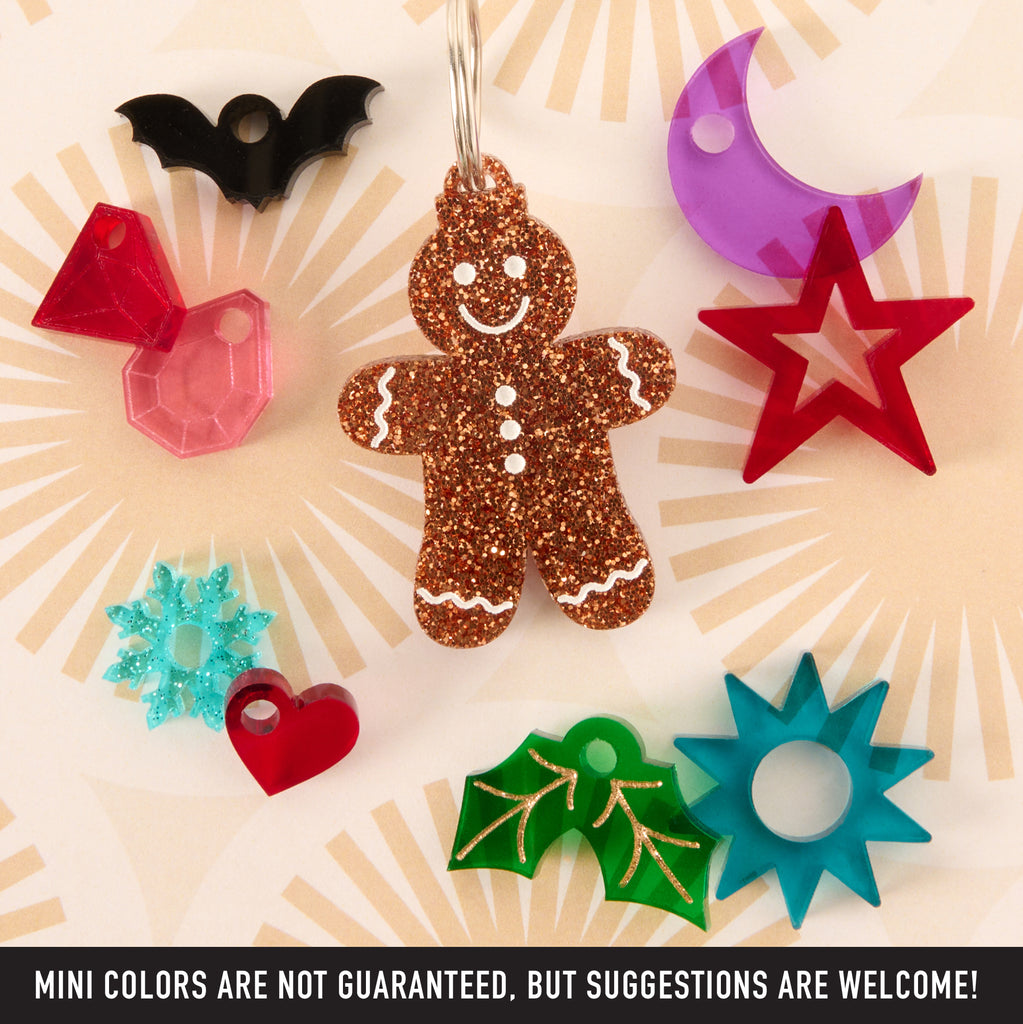 A central glitter gingerbread man charm is surrounded by assorted small acrylic charms in different colors and shapes: a black bat, pink gem, purple crescent moon, red star, teal snowflake, red heart, green holly, and blue sunburst/star shape. Text warns that "MINI COLORS ARE NOT GUARANTEED."