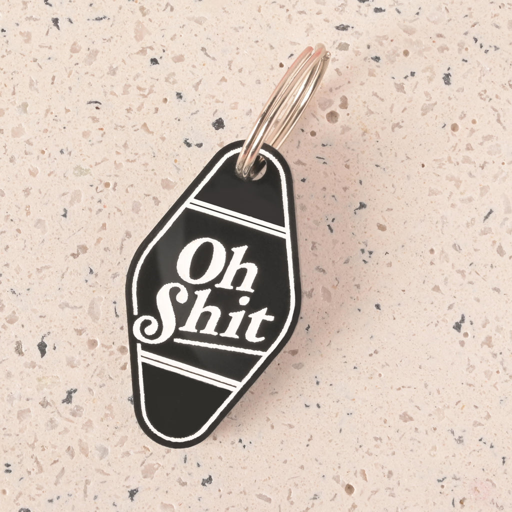 A close-up shot of a black hotel-style Dog and Cat tag that says "Oh Shit" in white letters. The keychain is attached to a silver ring and is resting on a light-colored, speckled surface.