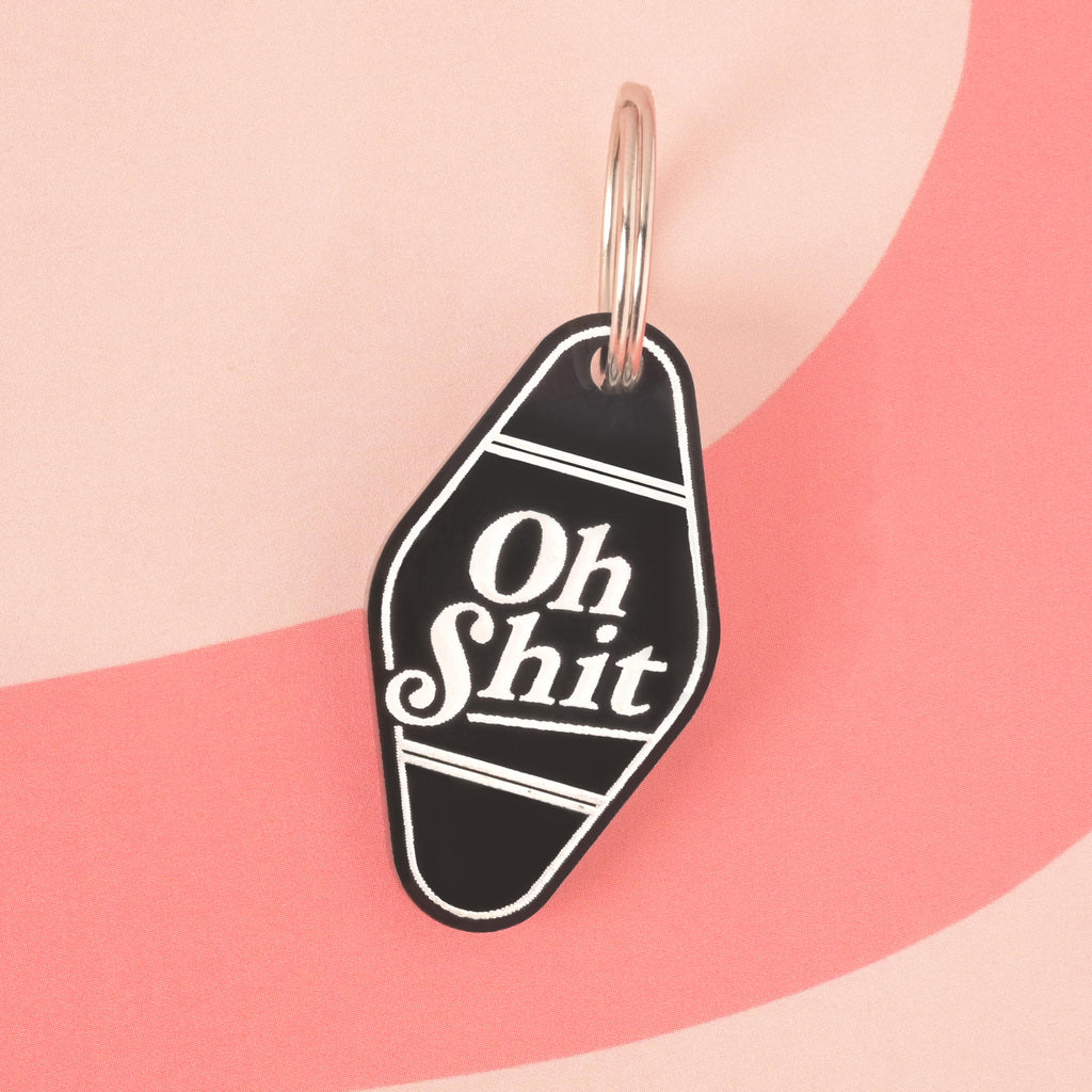 A black hotel-style Pet ID tag that says "Oh Shit" in white letters. The keychain is attached to a silver ring and is resting on a pink background with a wavy design.