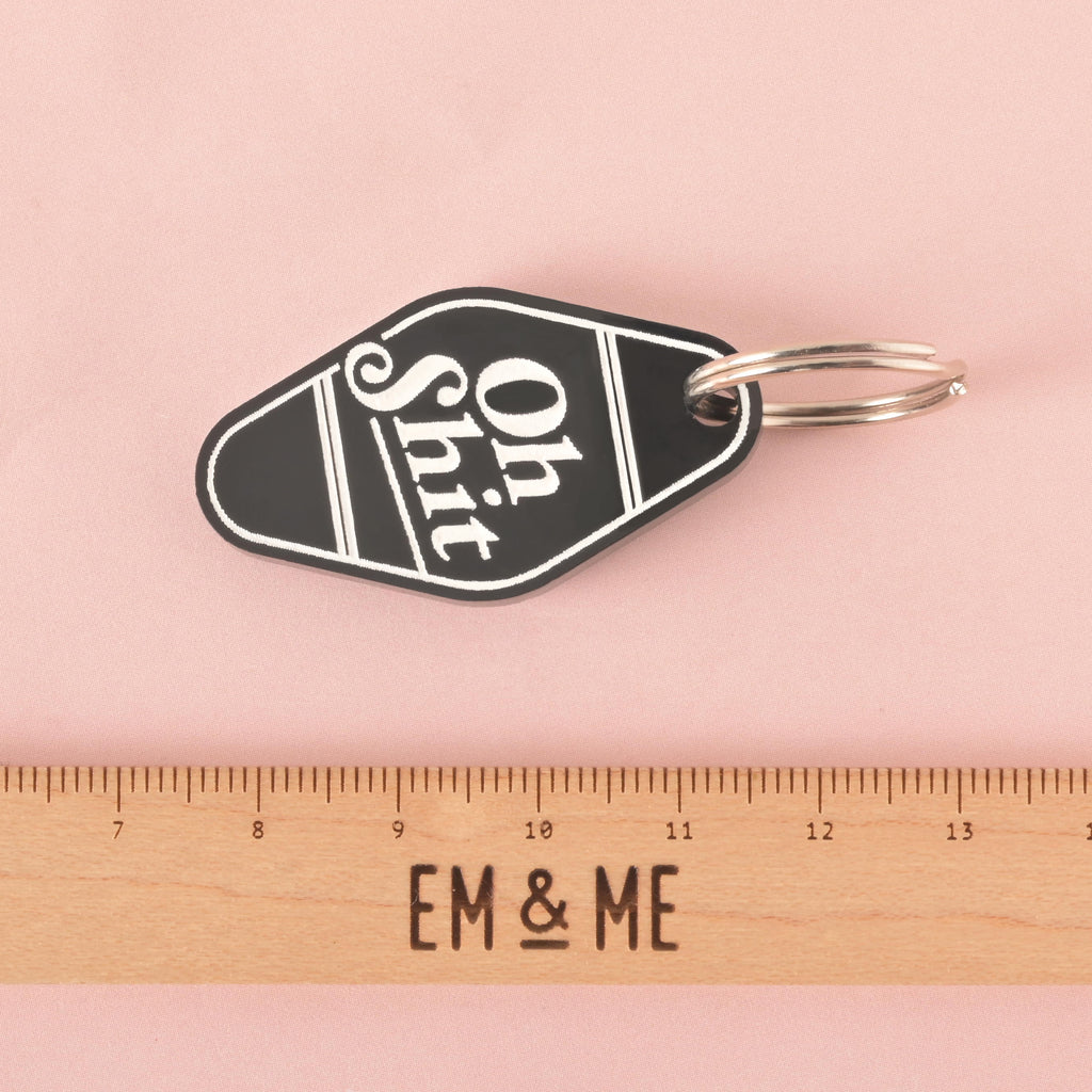 A black hotel-style Dog and Cat ID tag that says "Oh Shit" in white letters. A ruler with the "EM & ME" logo is placed below the keychain to show its size. The keychain is shown on a solid pink background.