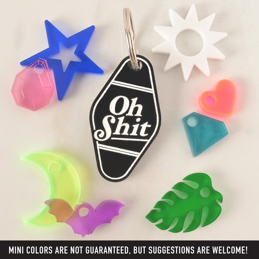A black hotel-style keychain that says "Oh Shit" in white letters. Surrounding it are various charms, including a blue star with a pink gemstone, a green monstera leaf, a white sun, a neon pink heart, a blue diamond, a neon yellow moon, and a purple bat. The image also contains the text "MINI COLORS ARE NOT GUARANTEED, BUT SUGGESTIONS ARE WELCOME!" The items are displayed on a white, speckled surface.
