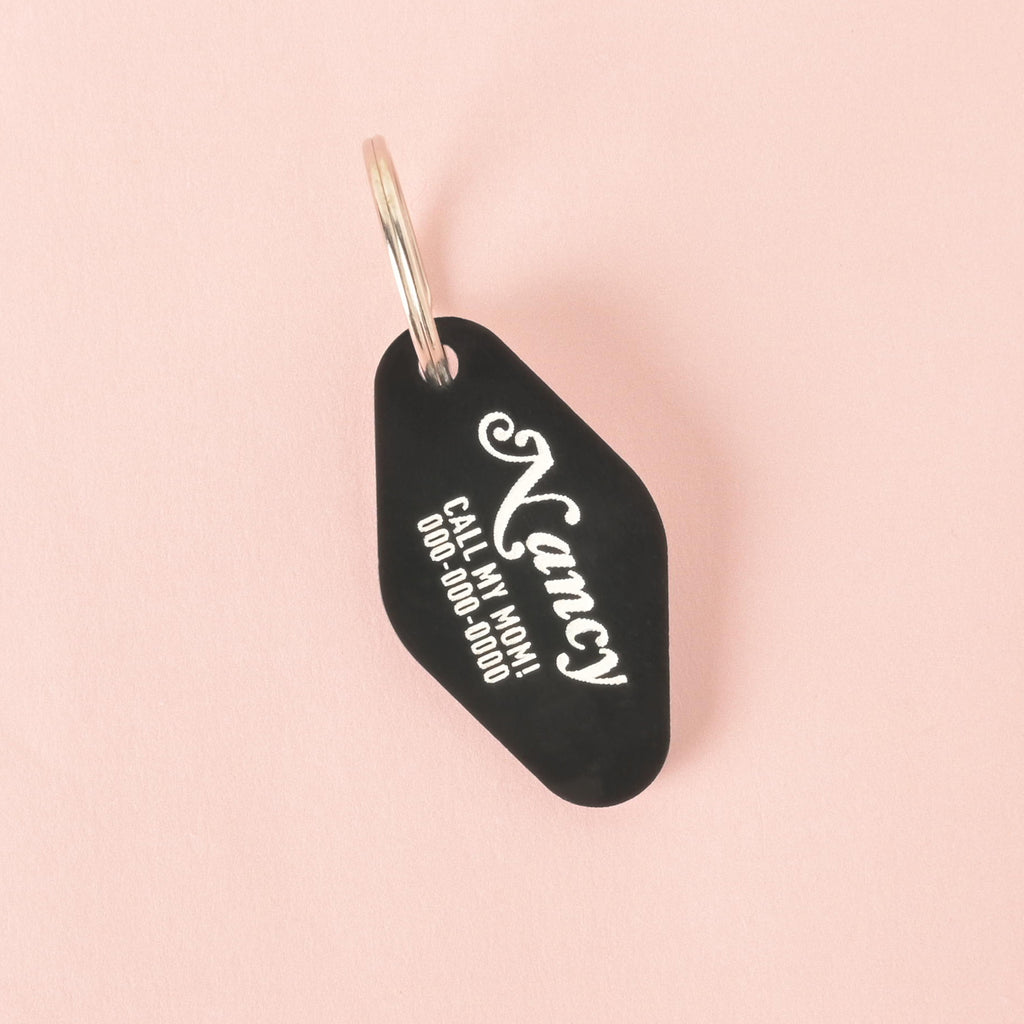 A close-up product photo of a black, retro motel-key-shaped pet tag. The tag is engraved with the name "Nancy" and the funny phrase "CALL MY MOM!" in white text, and is shown with a silver split key ring.