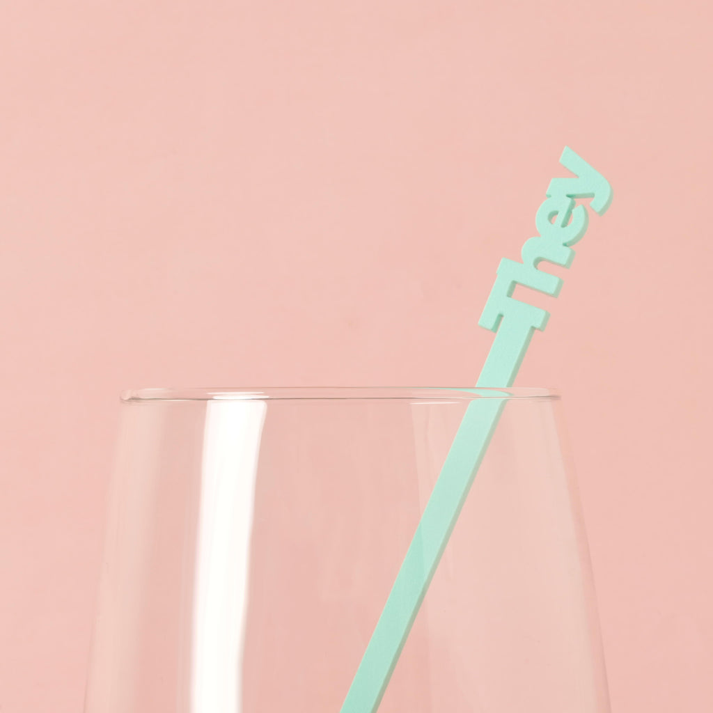 A single light blue cocktail stirrer with the word "they" is placed inside a clear glass. The background is a solid pink color.
