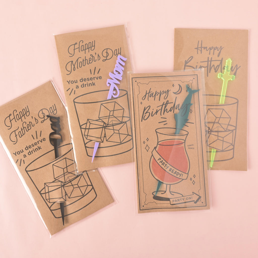 four different cocktail stirrer gift cards. The cards feature illustrations of a glass with a stirrer and a relevant holiday message, including "Happy Father's Day" with a black stirrer, "Happy Mother's Day" with a purple "Mom" stirrer, "Happy Birthday" with a teal swordfish stirrer, and "Happy Birthday" with a translucent green sword stirrer. The items are shown on a solid pink background.