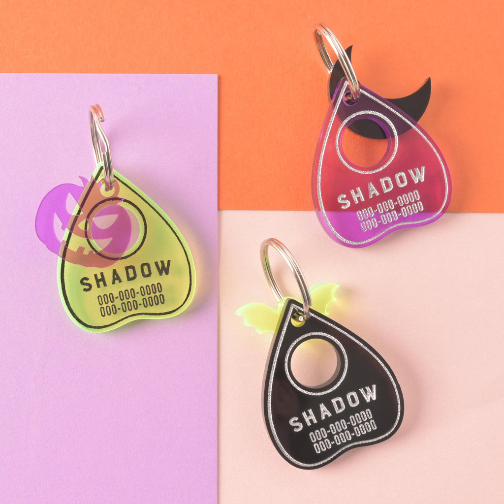 collection of three ouija planchet-shaped pet ID tags, One Fluorescent green with a purple jack-o-lantern mini charm, one purple with a black mini charm and the third black with a fluorescent green bat mini charm on a color blocked background.