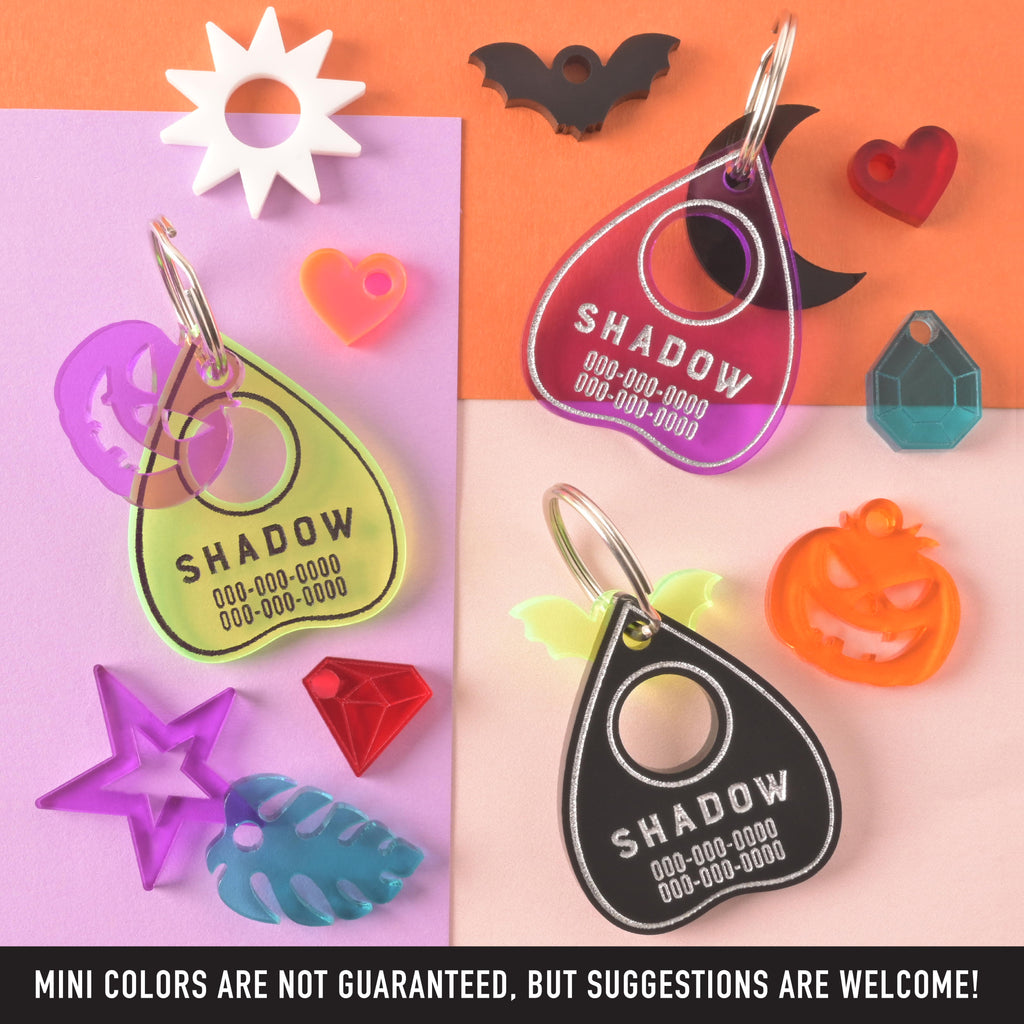 Collection of three ouija planchet-shaped pet tags surrounded by a selection of mini charms on color blocked background with a banner on the bottom that reads "MINI COLORS ARE NOT GUARANTEED, BUT SUGGESTIONS ARE WELCOME!"