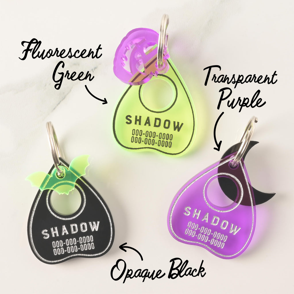 Collection of three ouija planchet-shaped personalized Pet ID tags. Fluorescent green tag with a purple jack-o-lantern mini, black pet tag with fluorescent green bat mini charm and a transparent purple pet tag with a black moon mini on marble background.