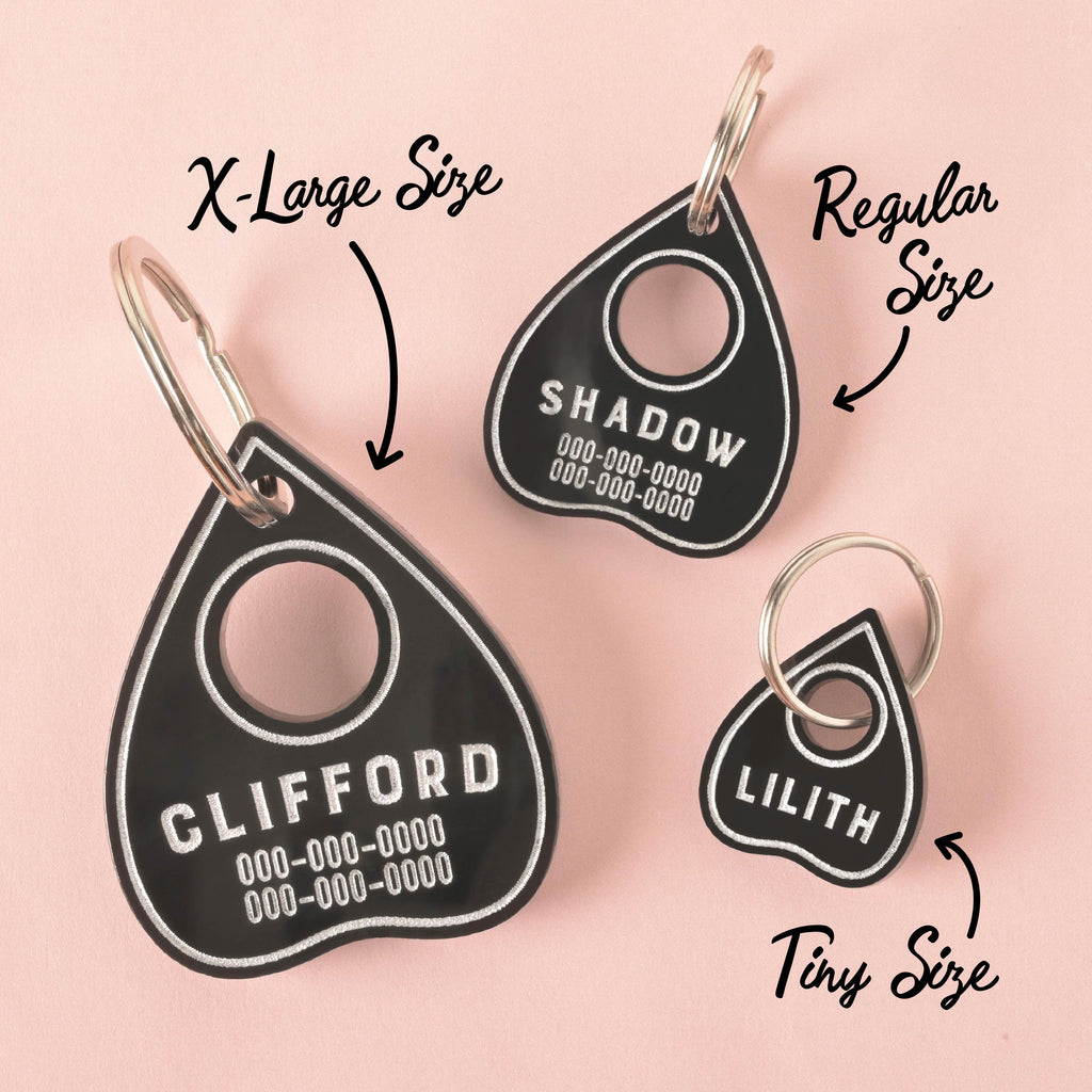 Three black Ouija planchette-shaped pet tags in different sizes. The largest tag, labeled "X-Large Size," has the name "CLIFFORD" on it. The middle tag, labeled "Regular Size," has the name "SHADOW," and the smallest tag, labeled "Tiny Size," has the name "LILITH." All tags are on a solid pink background.