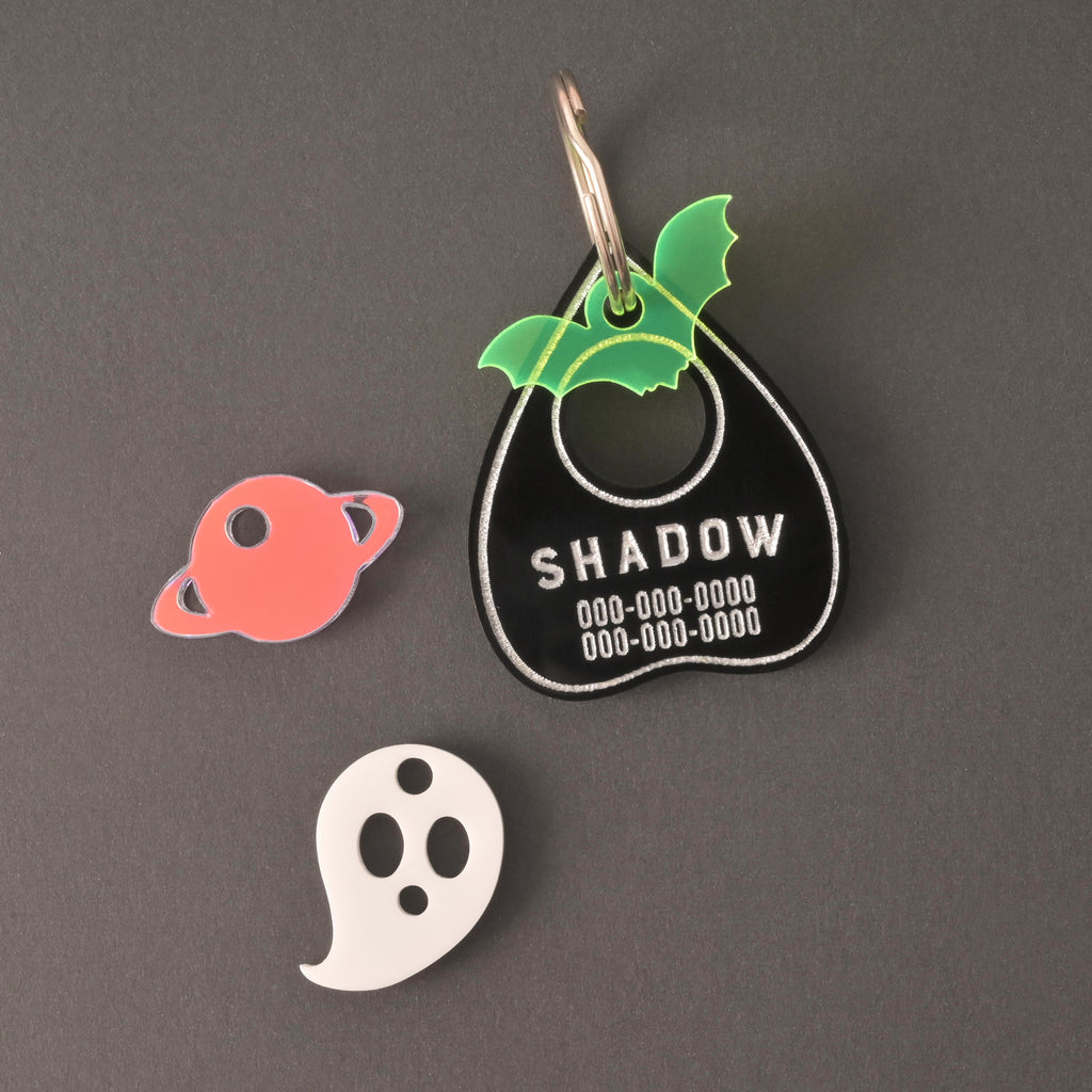 Black ouija planchet-shaped personalized pet tag with fluorescent green bat, iridescent Saturn and white ghost mini charms on solid black background.