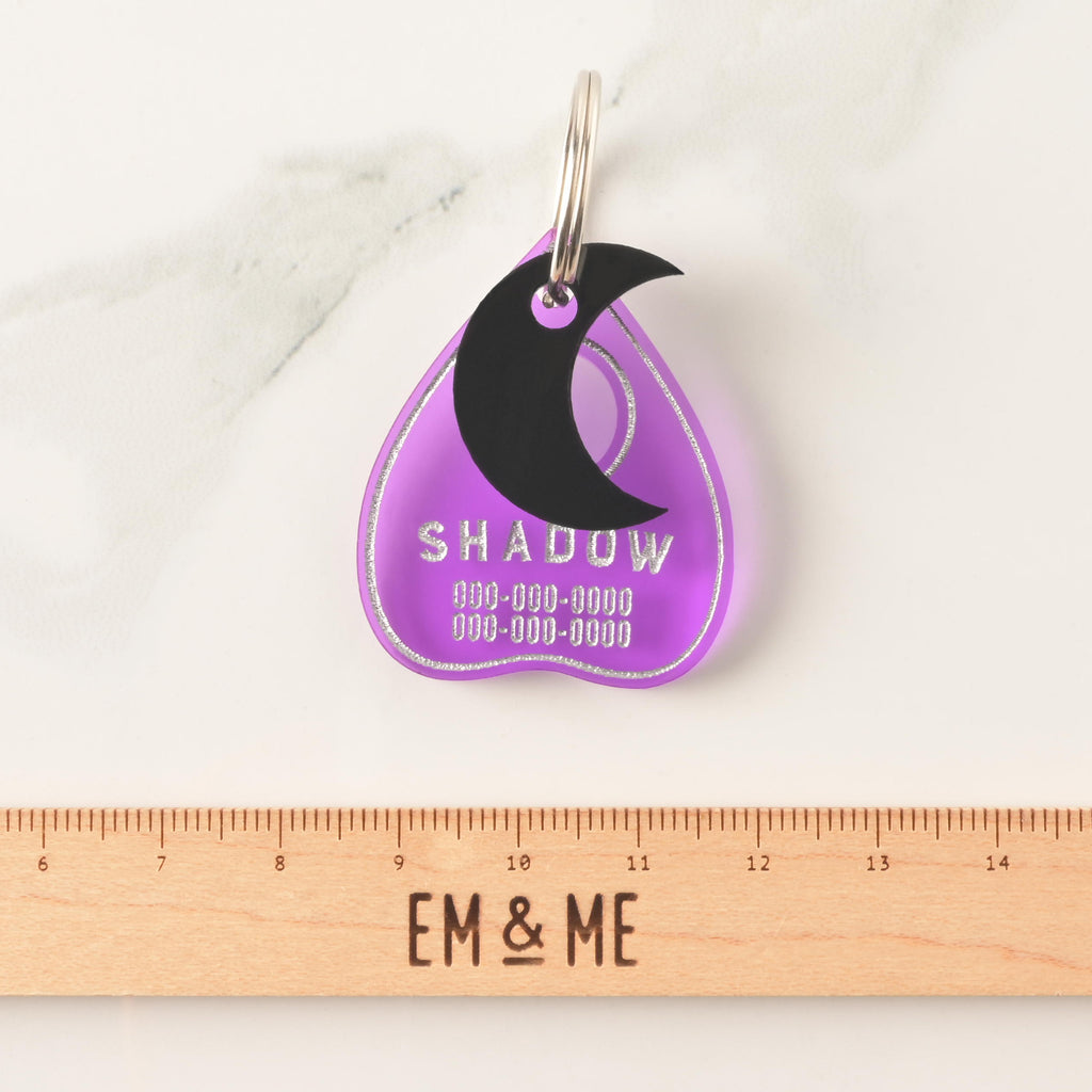 transparent purple ouija planchet0shaped pet tag with black moon mini charm on top of a wooden ruler that reads "EM & ME" on marble background.