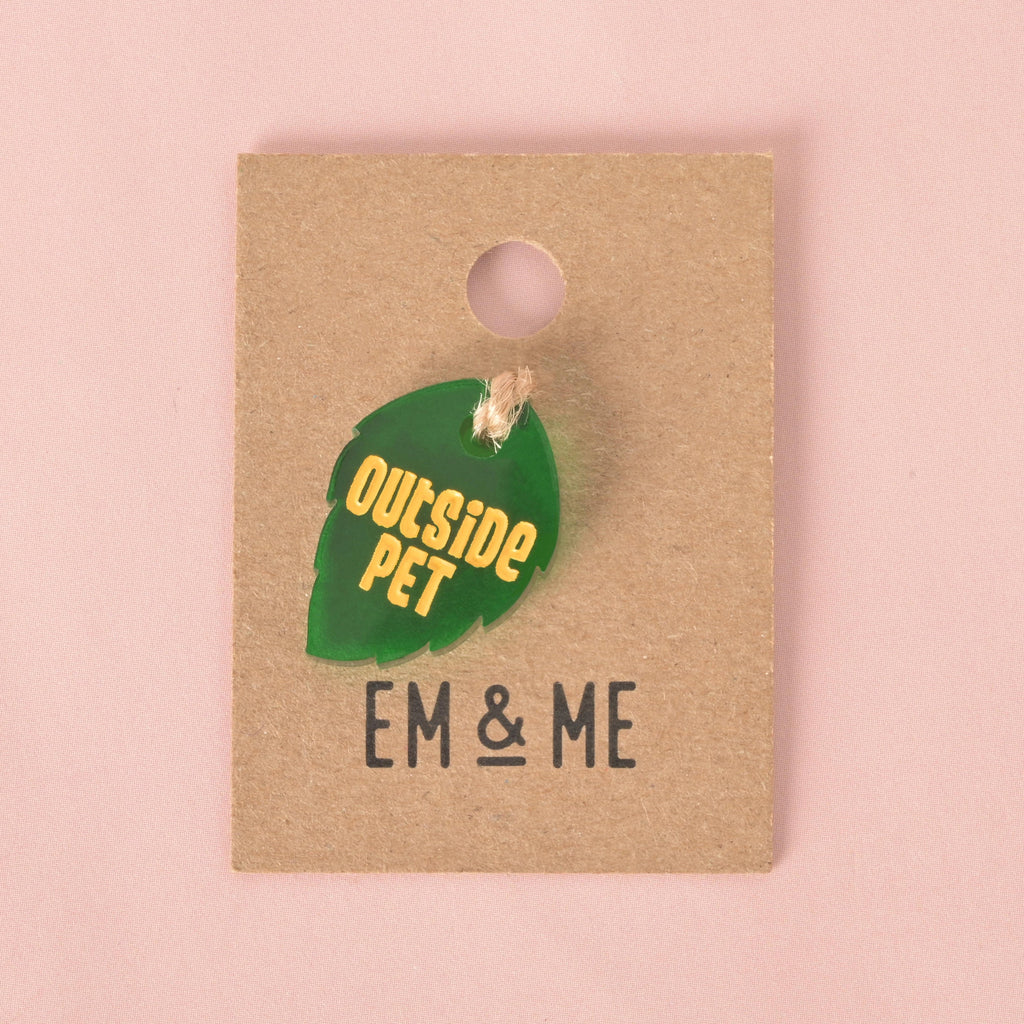 A small, dark green, leaf-shaped pet tag with serrated edges. The tag is engraved with the yellow text "OUTSIDE PET" and is attached with a piece of string to a brown cardboard tag with the logo "EM & ME." The item is displayed on a solid pink background.