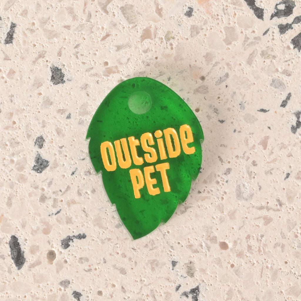 A close-up of a dark green, leaf-shaped pet tag with serrated edges. The tag is engraved with the yellow text "OUTSIDE PET" and is displayed on a light-colored, speckled countertop or stone surface.