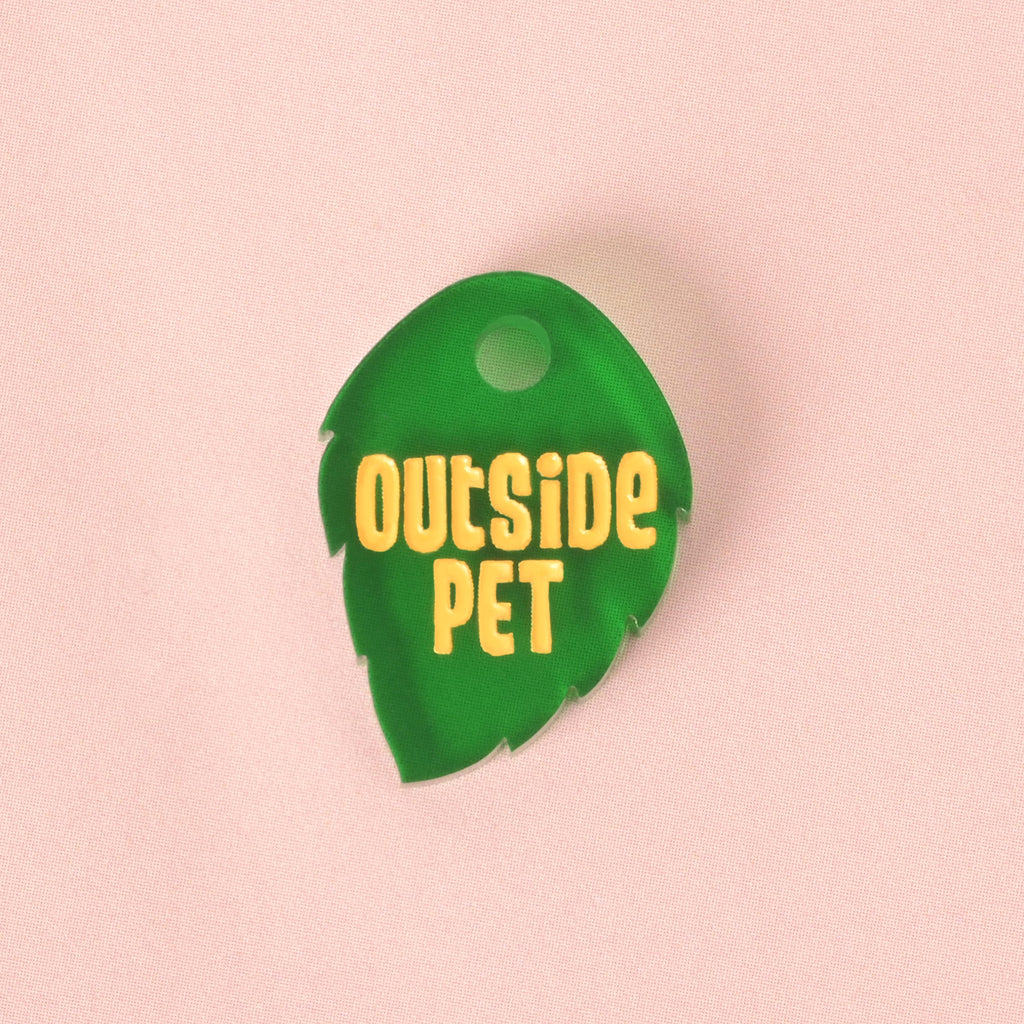 A close-up of a dark green, leaf-shaped pet tag with serrated edges. The tag is engraved with the yellow text "OUTSIDE PET" and is displayed on a solid pink background.