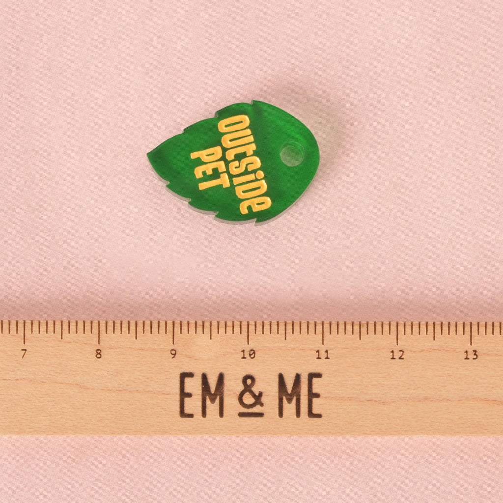 A small, dark green, leaf-shaped pet tag with serrated edges. The tag is engraved with the yellow text "OUTSIDE PET." It is displayed above a wooden ruler with the logo "EM & ME" on a solid pink background.