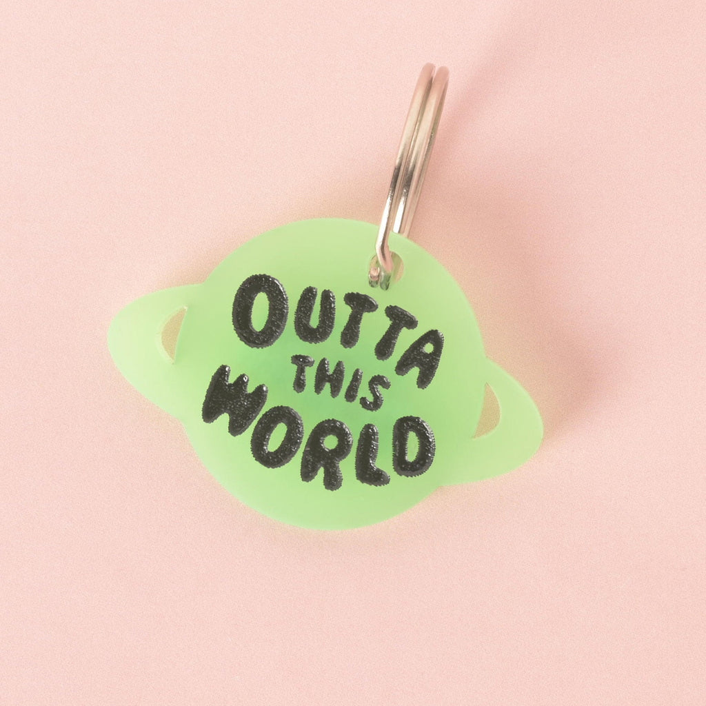 A close-up of a custom, glow-in-the-dark, planet-shaped pet ID tag with a shiny silver keyring. The tag is engraved with the black text "OUTTA THIS WORLD." The tag is displayed on a solid pink background.