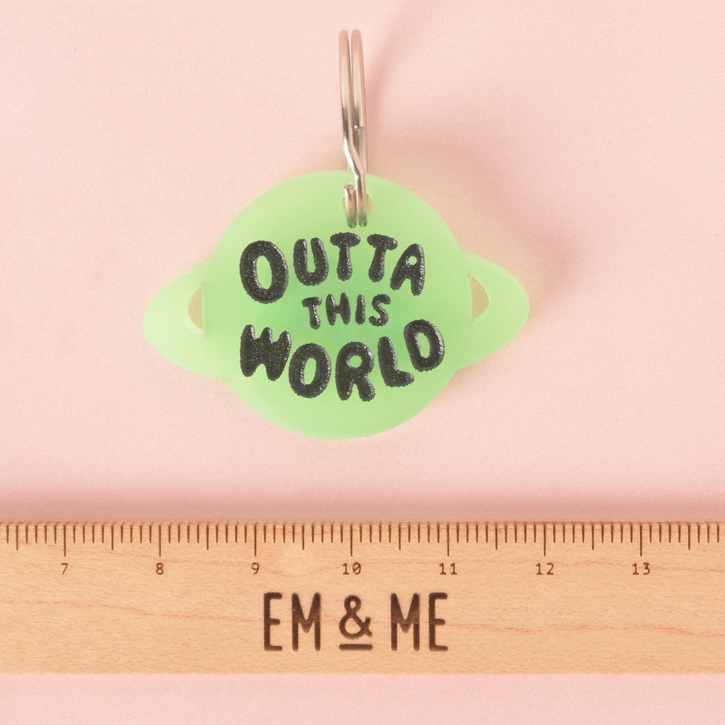 A custom, glow-in-the-dark, planet-shaped pet ID tag with a black engraving that says "OUTTA THIS WORLD." The tag is attached to a silver keyring. A wooden ruler with the logo "EM & ME" is visible at the bottom of the photo, which is set on a solid pink background.