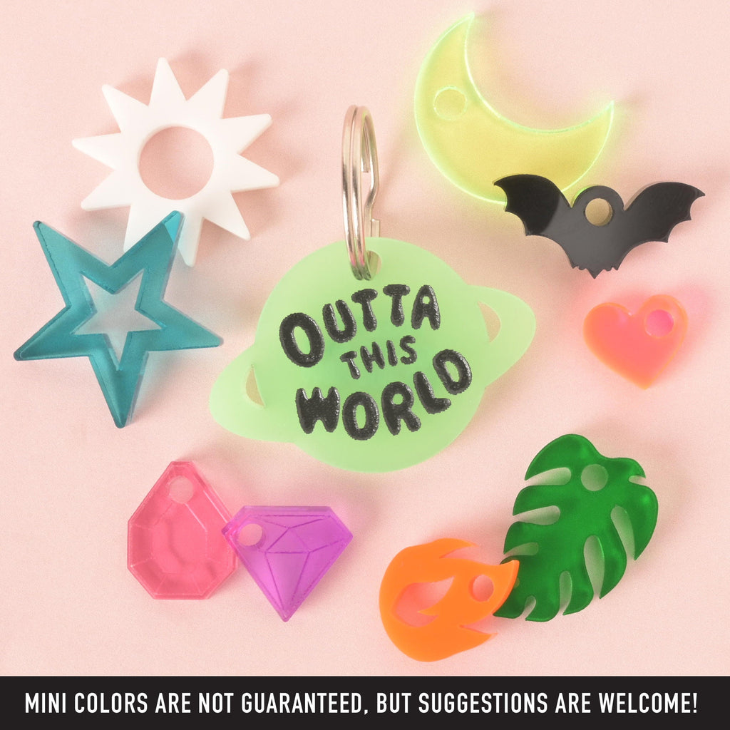 A custom, glow-in-the-dark, planet-shaped pet ID tag with a black engraving that says "OUTTA THIS WORLD." The tag is surrounded by a variety of mini charms, including a white sunburst, a translucent blue star, a pink gem, a purple gem, an orange flame, a green tropical leaf, a pink heart, a black bat, and a glow-in-the-dark crescent moon. The items are arranged on a pink background. A note at the bottom says, "MINI COLORS ARE NOT GUARANTEED, BUT SUGGESTIONS ARE WELCOME!"