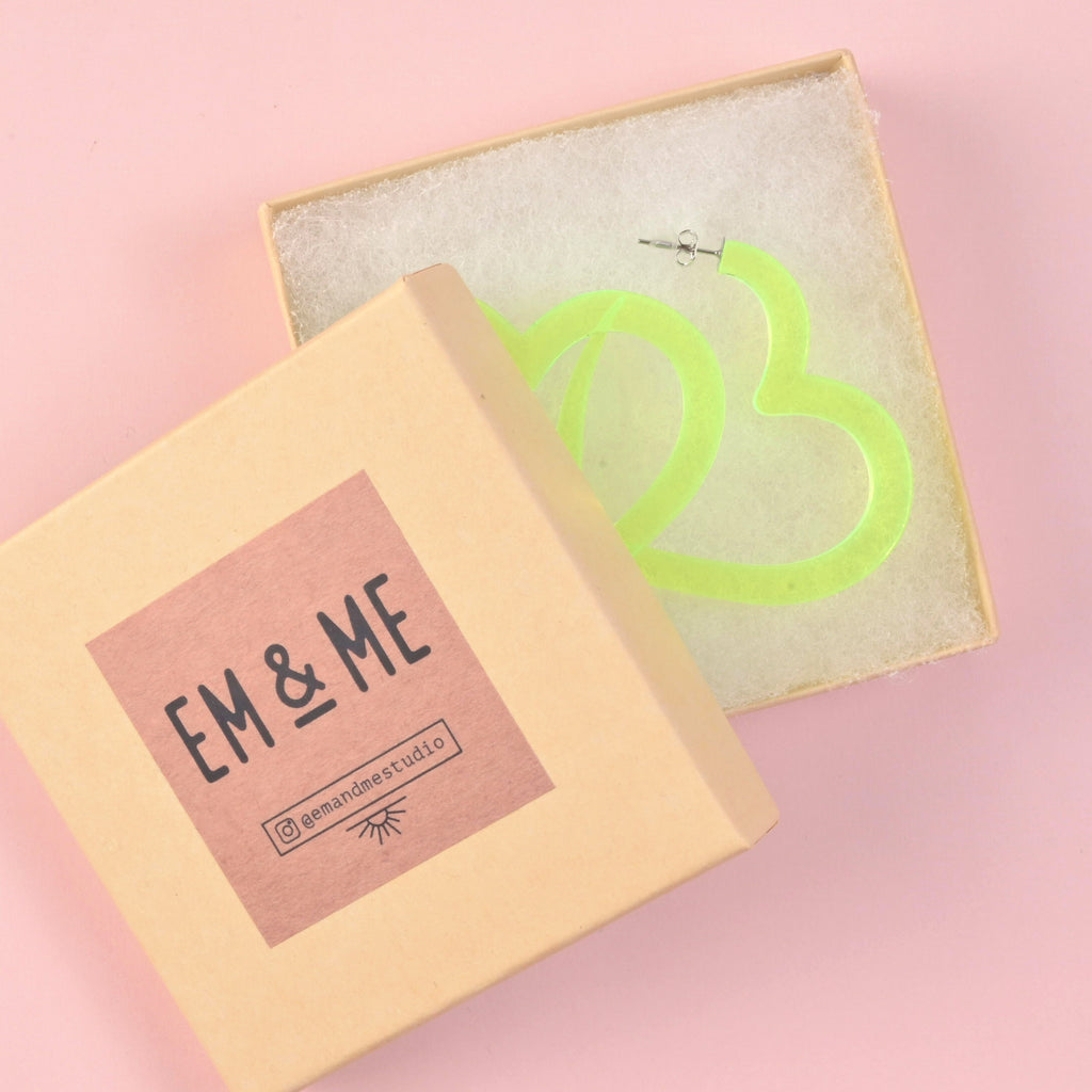 A pair of oversized fluorescent neon green acrylic heart hoop earrings lying inside a white-lined gift box. The lid, branded with 'EM & ME,' rests in the foreground, indicating they are ready to gift.