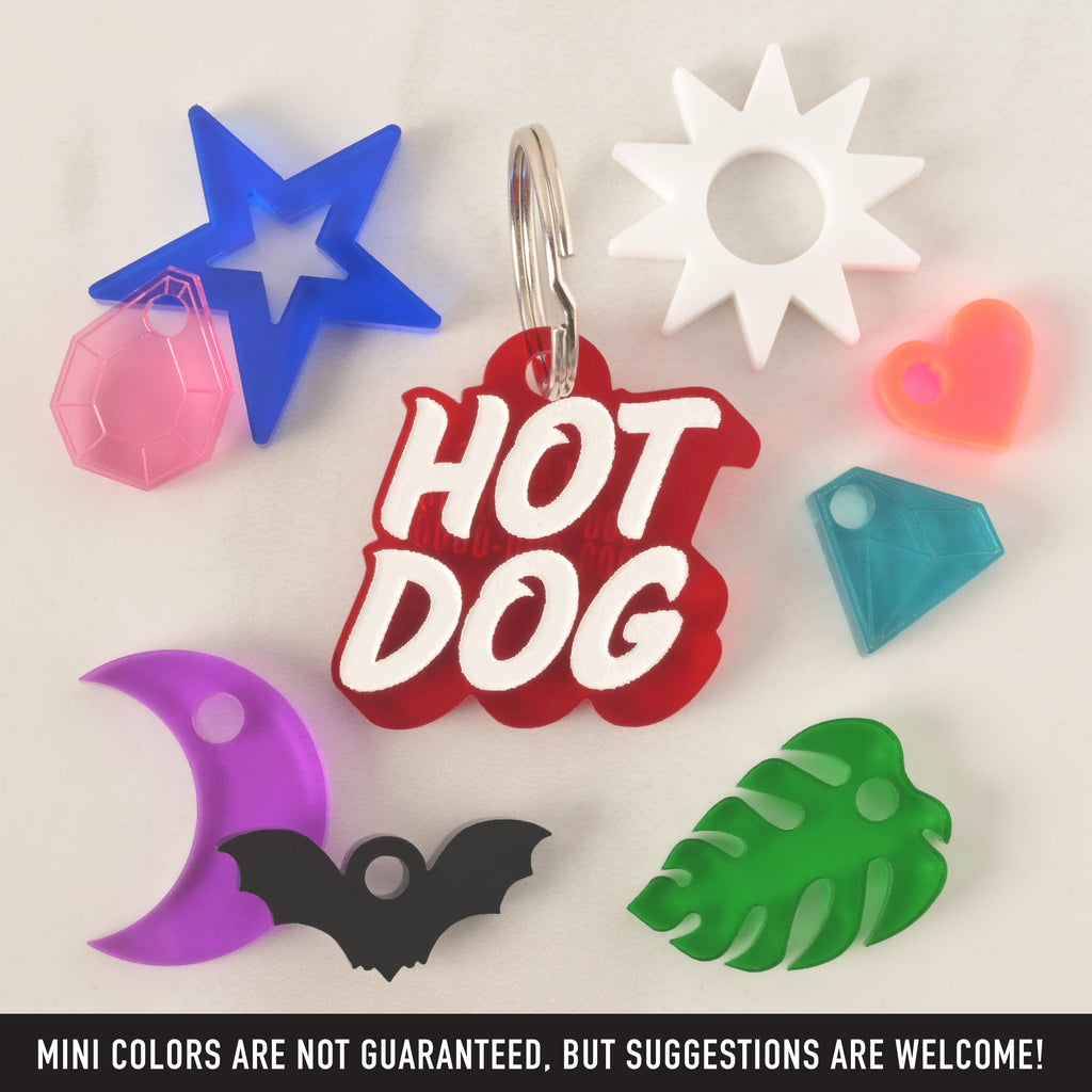 A collection of pet tags and charms scattered on a white surface. The central piece is a red "HOT DOG" tag in a groovy font. Surrounding it are various charms, including a blue star with a pink gemstone, a purple crescent moon, a black bat, a green monstera leaf, a white sun, a neon pink heart, and a blue diamond.