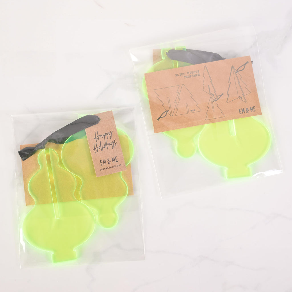 Two packaged sets of flat-pack fluorescent green icicle-shaped Christmas tree ornaments. The package on the left contains two ornaments and has a "Happy Holidays" tag attached with a dark ribbon. The package on the right shows three icicle pieces that slide together to form a 3D ornament. Both packages have "EM & ME" logos and assembly instructions. The items are displayed on a white marble surface.