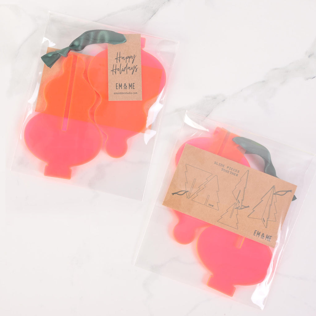 Two packaged sets of flat-pack icicle-shaped Christmas tree ornaments in neon pink and orange. The package on the left contains two ornaments and has a "Happy Holidays" tag attached with a dark green ribbon. The package on the right shows three icicle pieces that slide together to form a 3D ornament. Both packages have "EM & ME" logos. The items are displayed on a white marble surface.