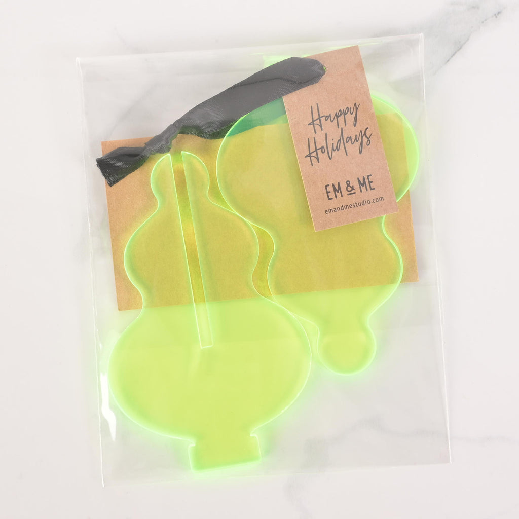 A packaged set of flat-pack fluorescent green icicle-shaped Christmas tree ornaments. The package contains two ornaments and has a "Happy Holidays" tag attached with a dark ribbon. The tag also includes the "EM & ME" logo. The items are displayed on a white marble surface.